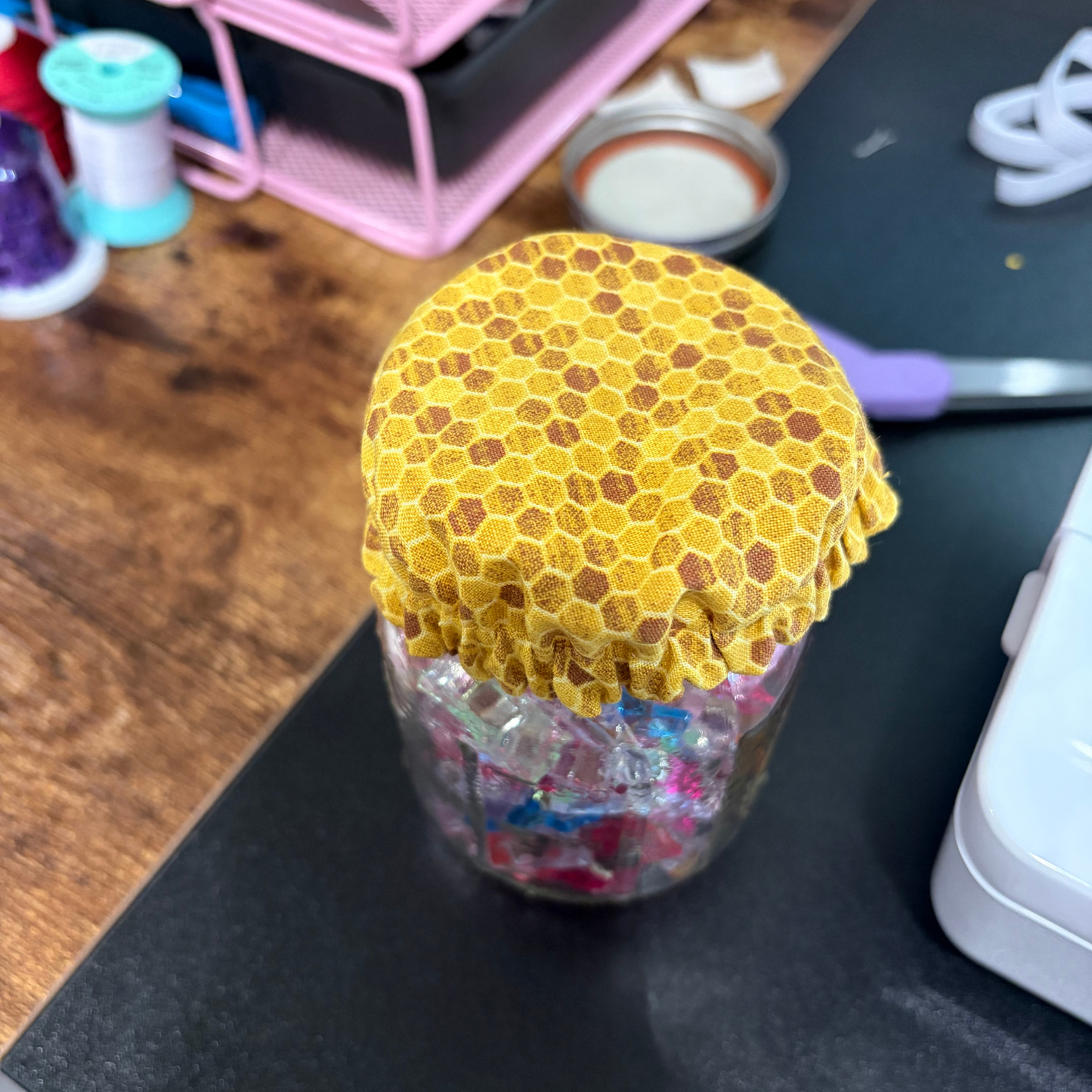 A clear glass jar filled with colorful items, covered with a yellow honeycomb-patterned fabric lid, set on a black desk with sewing supplies in the background.