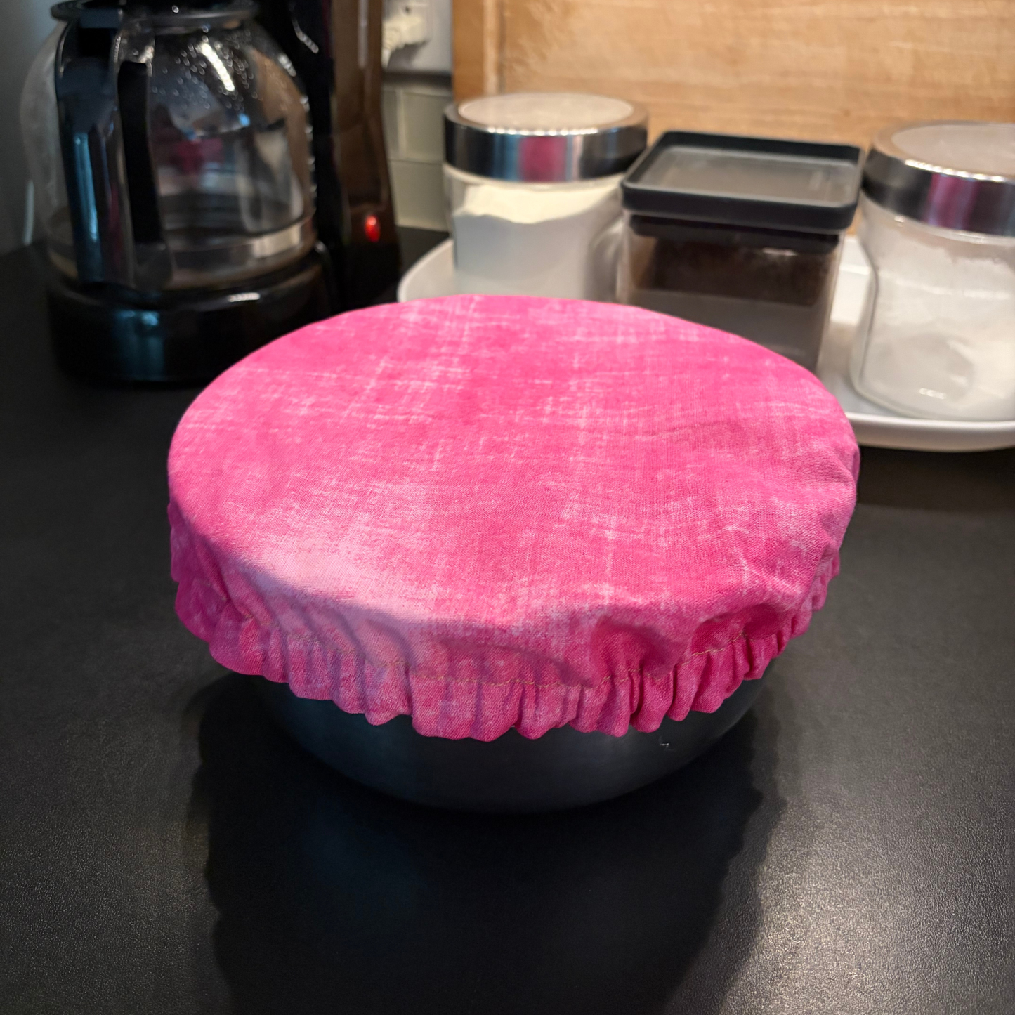 A bright pink fabric bowl cover with a gathered ruffled edge, shown draped over a round container on a dark kitchen countertop with a coffee maker and jars in the background.