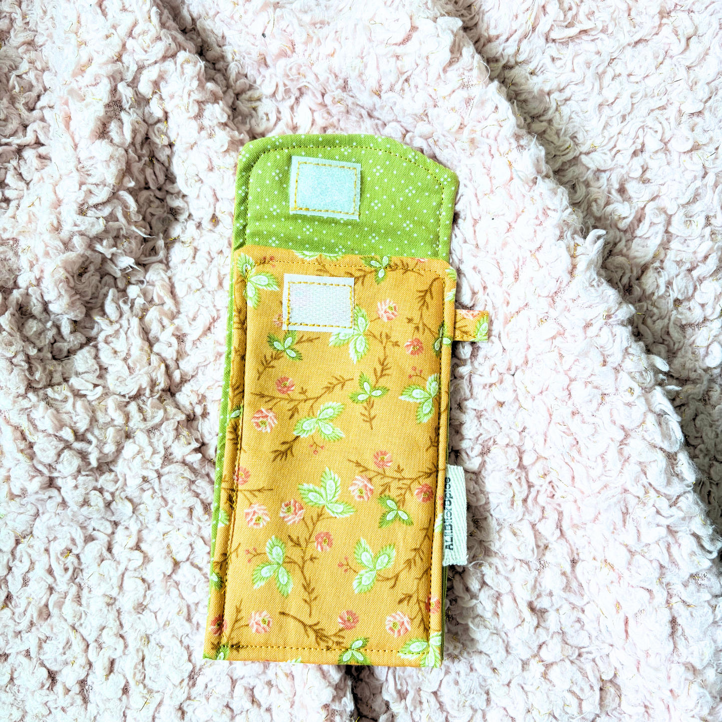 A handmade fabric sunglasses case with a green floral print and green trim, laid flat on a textured cream sherpa blanket.