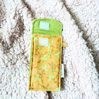 A handmade fabric sunglasses case with a green floral print and green trim, laid flat on a textured cream sherpa blanket.