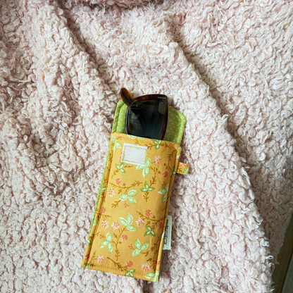 An orange floral fabric sunglasses pouch with a green interior lining, holding a pair of sunglasses, laid flat on a textured pink fleece blanket.