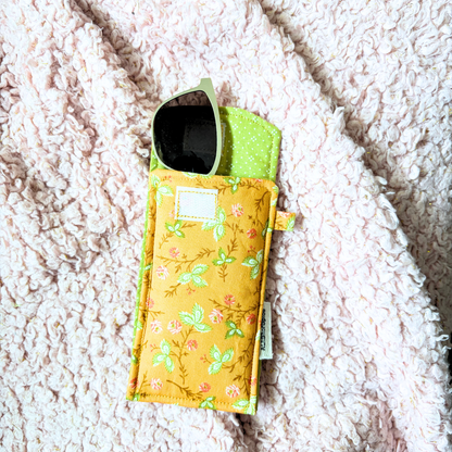 A fabric sunglasses pouch with a green and peach floral print and a lime green polka-dot interior, holding a pair of sunglasses, laid flat on a textured pink blanket.