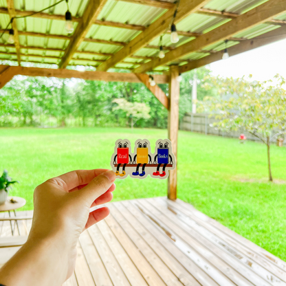 Hand holding a vinyl sticker with three book characters with the text ‘Read Banned Books’ with a green lawn background