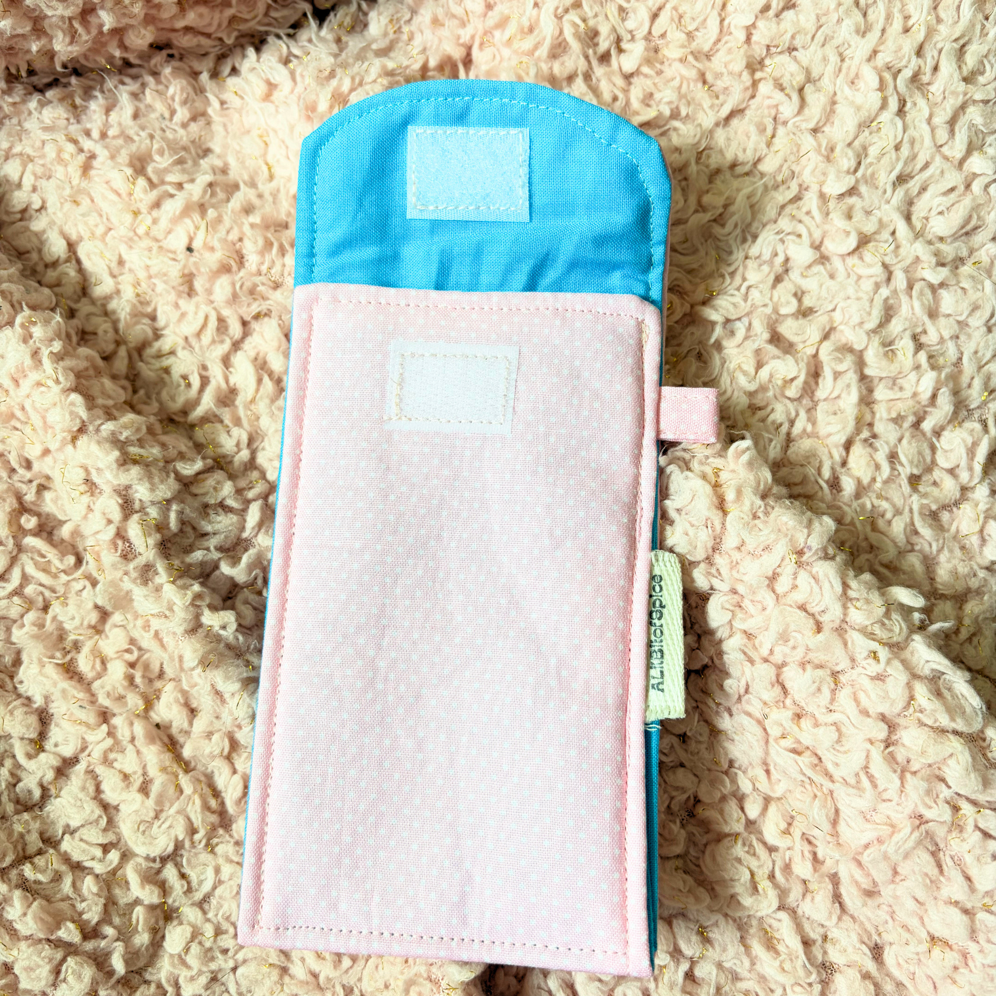 A pastel pink and light blue fabric sunglasses case with a small white label patch, laid flat on a textured cream sherpa background.