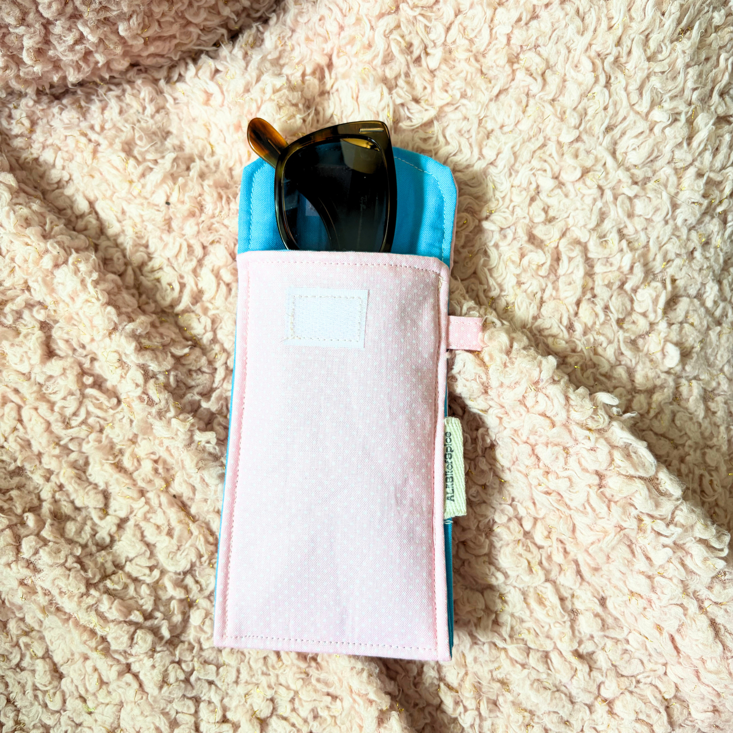A light pink fabric sunglasses pouch with a small white label on the side, shown on a textured cream sherpa background.