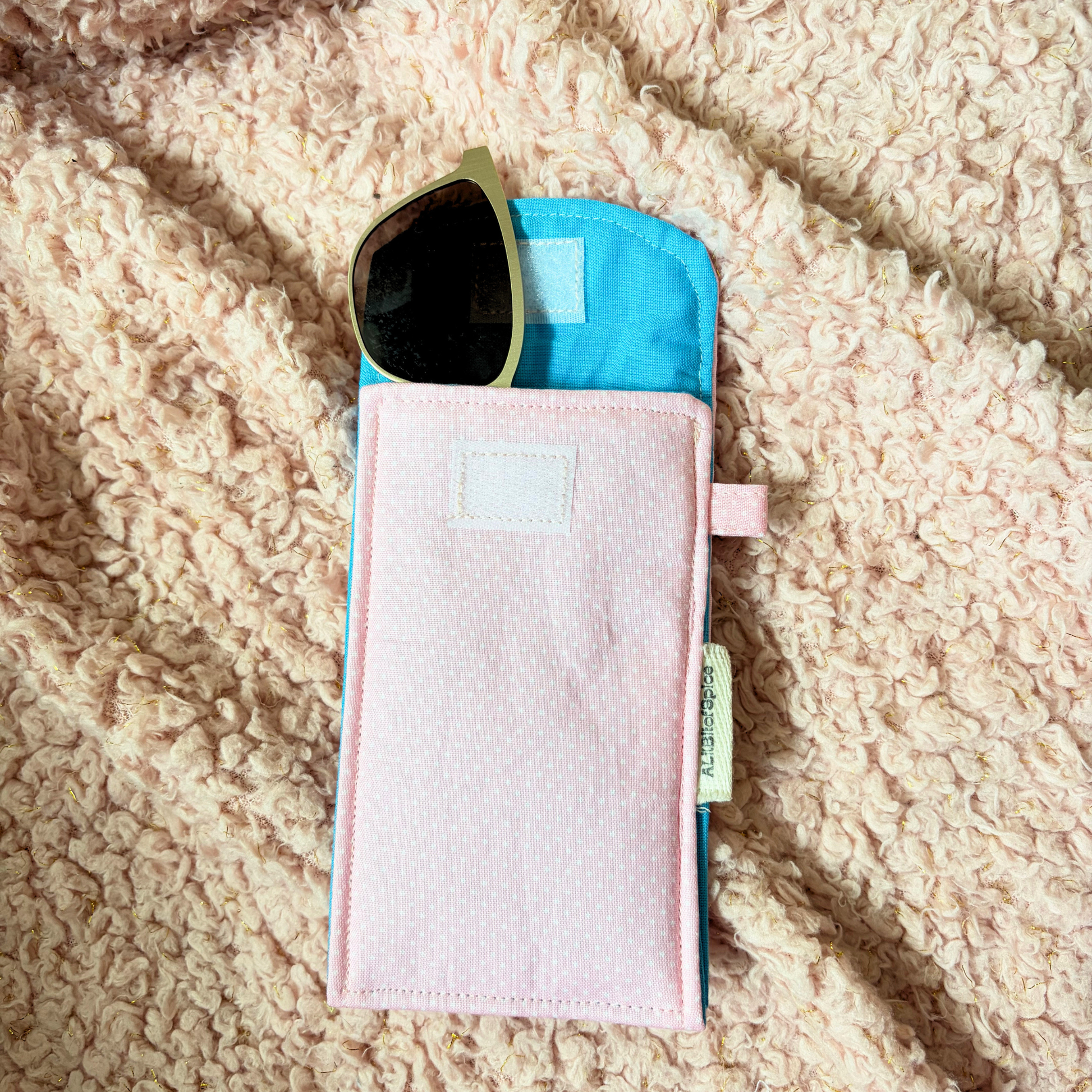 A light pink sunglasses pouch with a bright blue lining, shown on a beige sherpa blanket with sunglasses tucked inside.