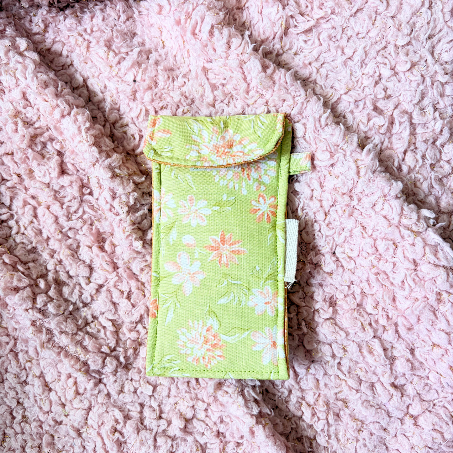 A small fabric pouch with a light green floral print, featuring white and peach flowers, laid flat on a pink textured fleece blanket.