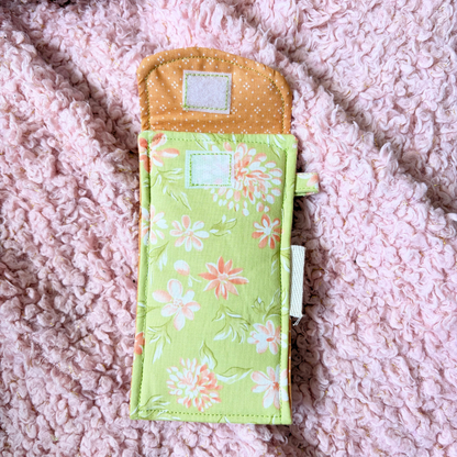 A handmade fabric pouch with a light green and orange floral print and a contrasting orange polka-dot top flap, displayed on a pink textured blanket.