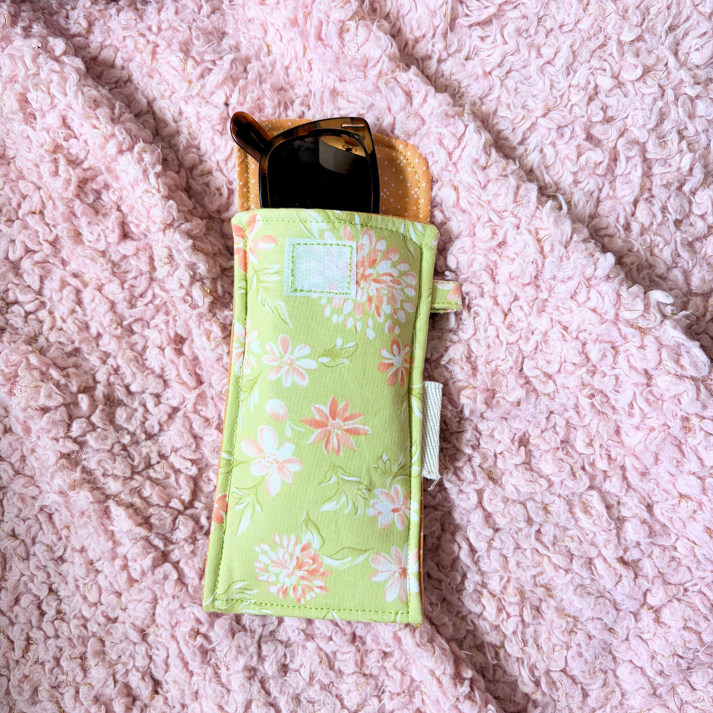 A light green fabric sunglasses case with a floral print, holding a pair of sunglasses, laid flat on a pink textured blanket.