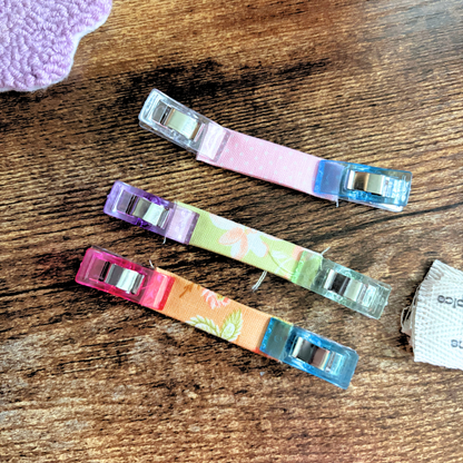 Four small plastic clips with colorful fabric straps are arranged diagonally on a wooden surface. The clips have pink, purple, orange, and blue translucent ends, and the center straps feature pastel floral and leaf patterns.