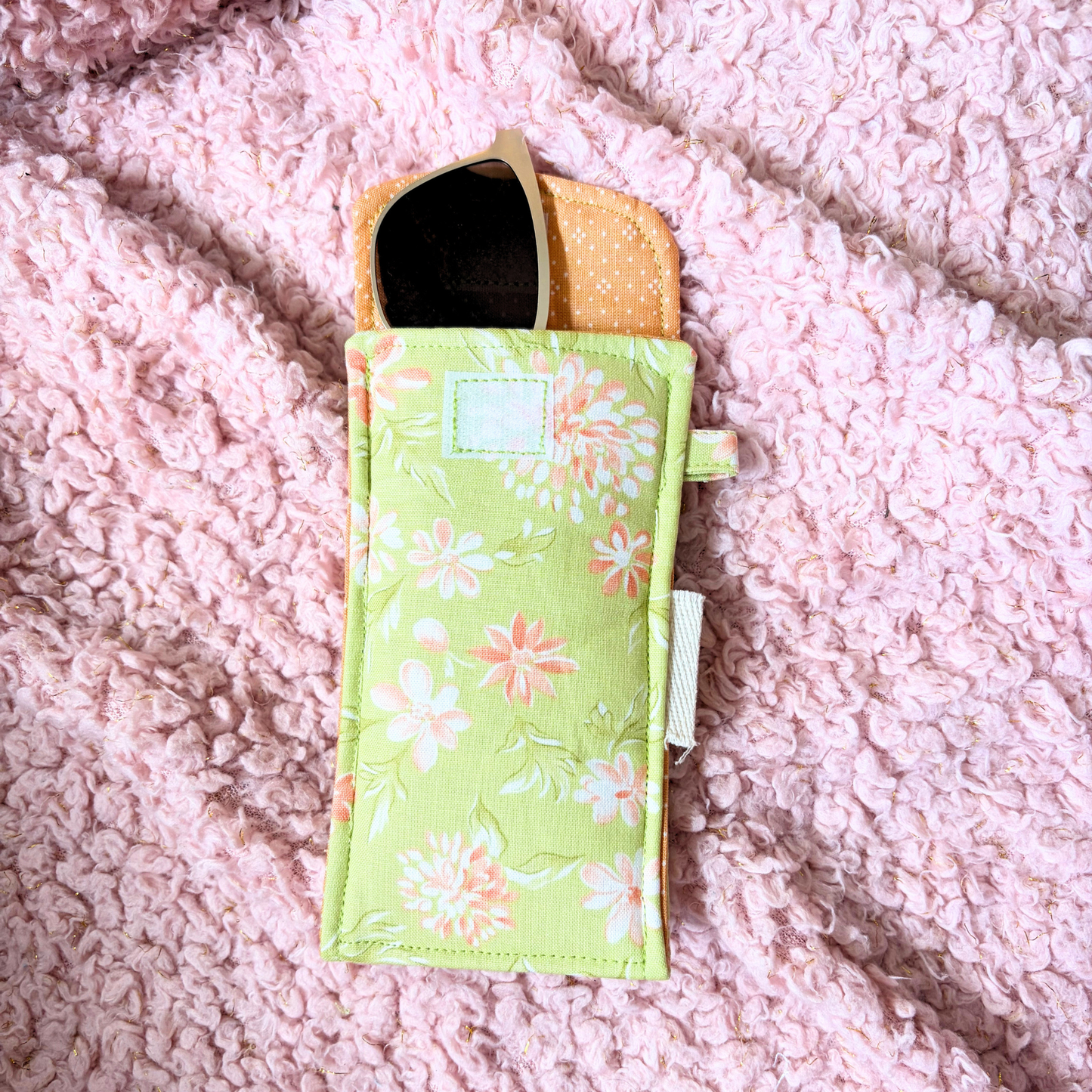 A light green fabric phone pouch with a white and orange floral print, shown open on a textured pink blanket background.