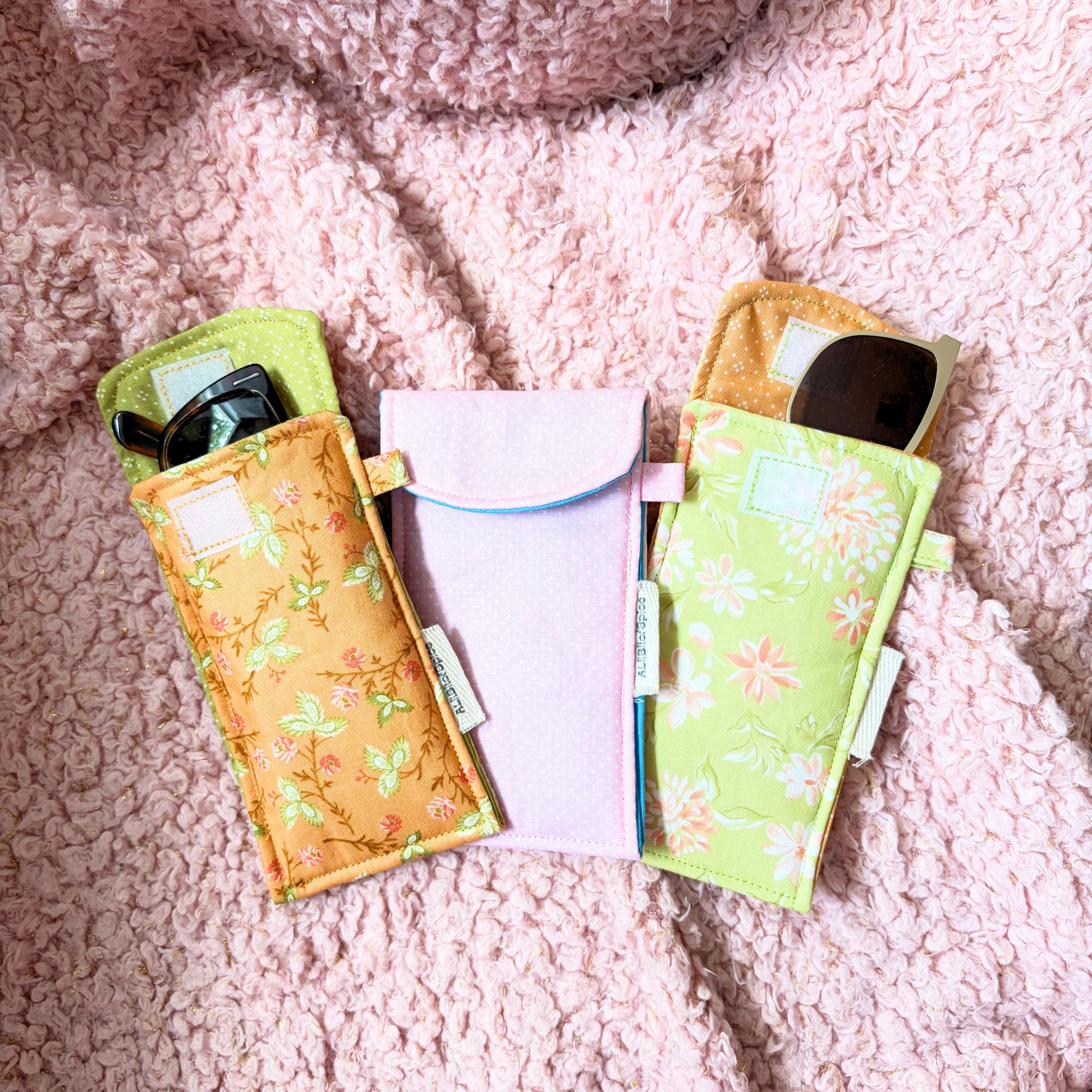 Three fabric sunglasses cases arranged on a pink textured blanket. Two cases have floral prints in yellow-orange and light green, and one center case is polka-dot light pink.