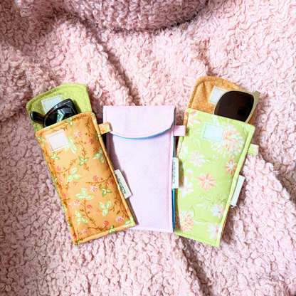 Three fabric sunglasses cases arranged on a pink textured blanket. Two cases have floral prints in yellow-orange and light green, and one center case is polka-dot light pink.