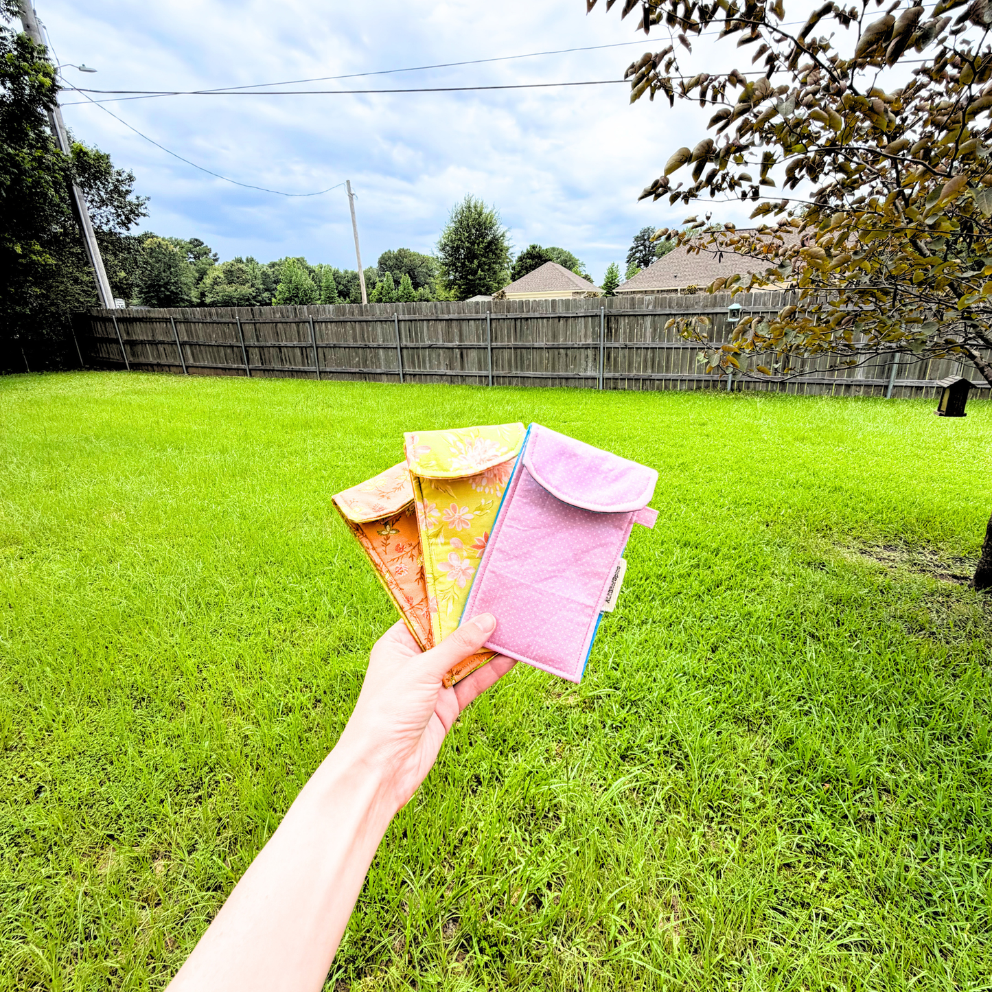 A hand holds three small colorful sunglasses cases in front of a grassy backyard with a wooden fence and trees.