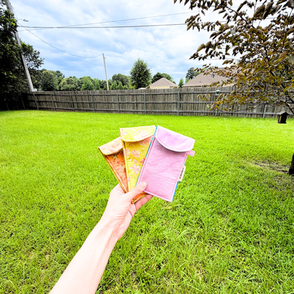 A hand holds three small colorful sunglasses cases in front of a grassy backyard with a wooden fence and trees.