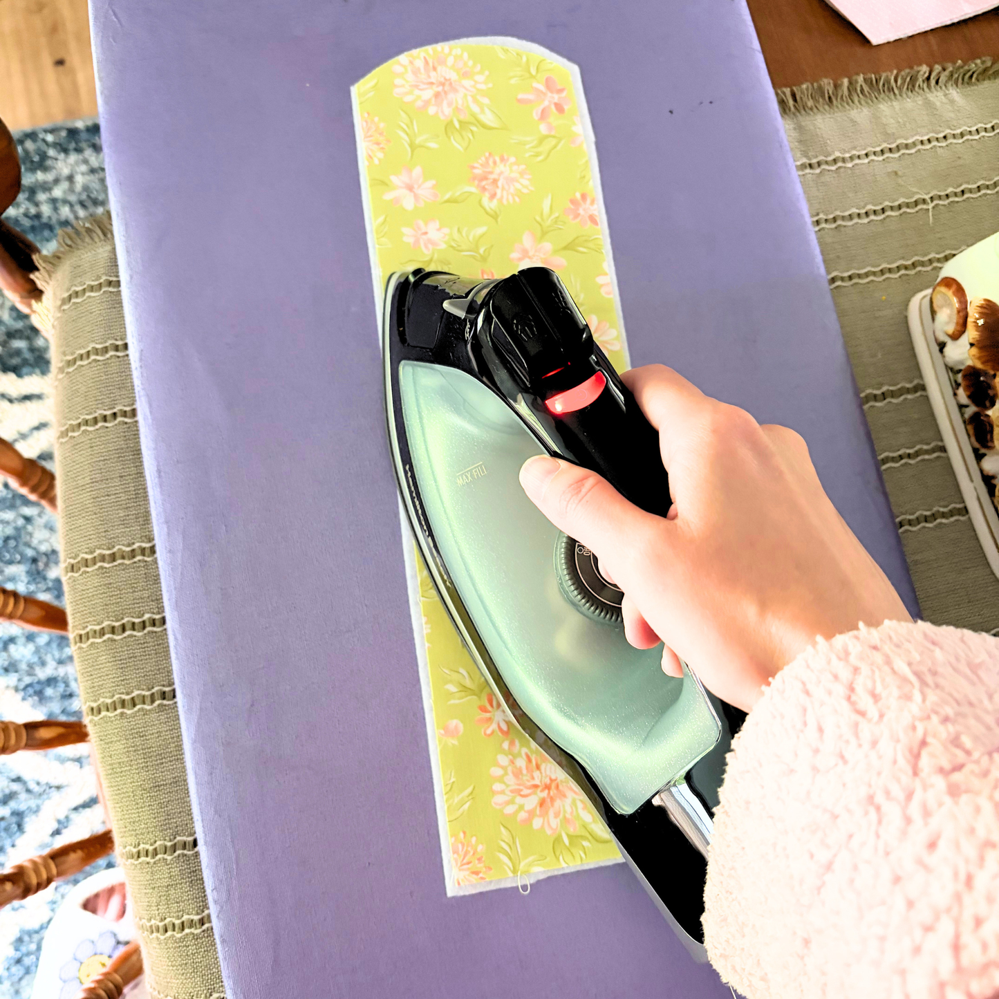 A hand ironing a piece of light green floral-patterned fabric on a lavender ironing board, with a black and mint green steam iron visible.