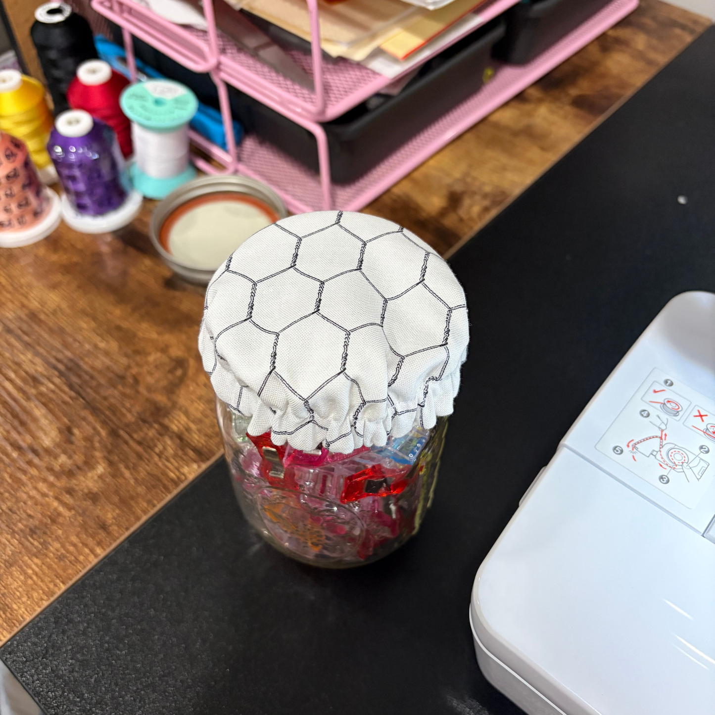 A clear glass jar filled with colorful craft pieces, covered with a white fabric lid featuring a black chicken wire pattern, sitting on a black tabletop next to a white machine.