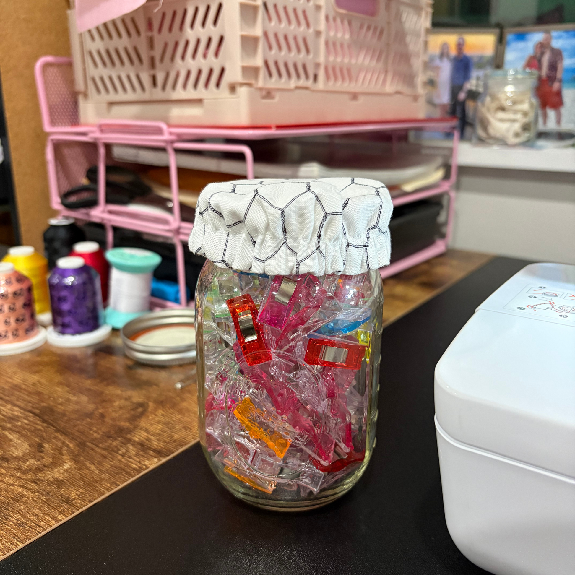 A clear glass jar filled with colorful translucent plastic pieces and covered with a white chicken wire patterned cloth lid, sitting on a dark tabletop in a craft or sewing room with spools of thread and a pink storage rack in the background.