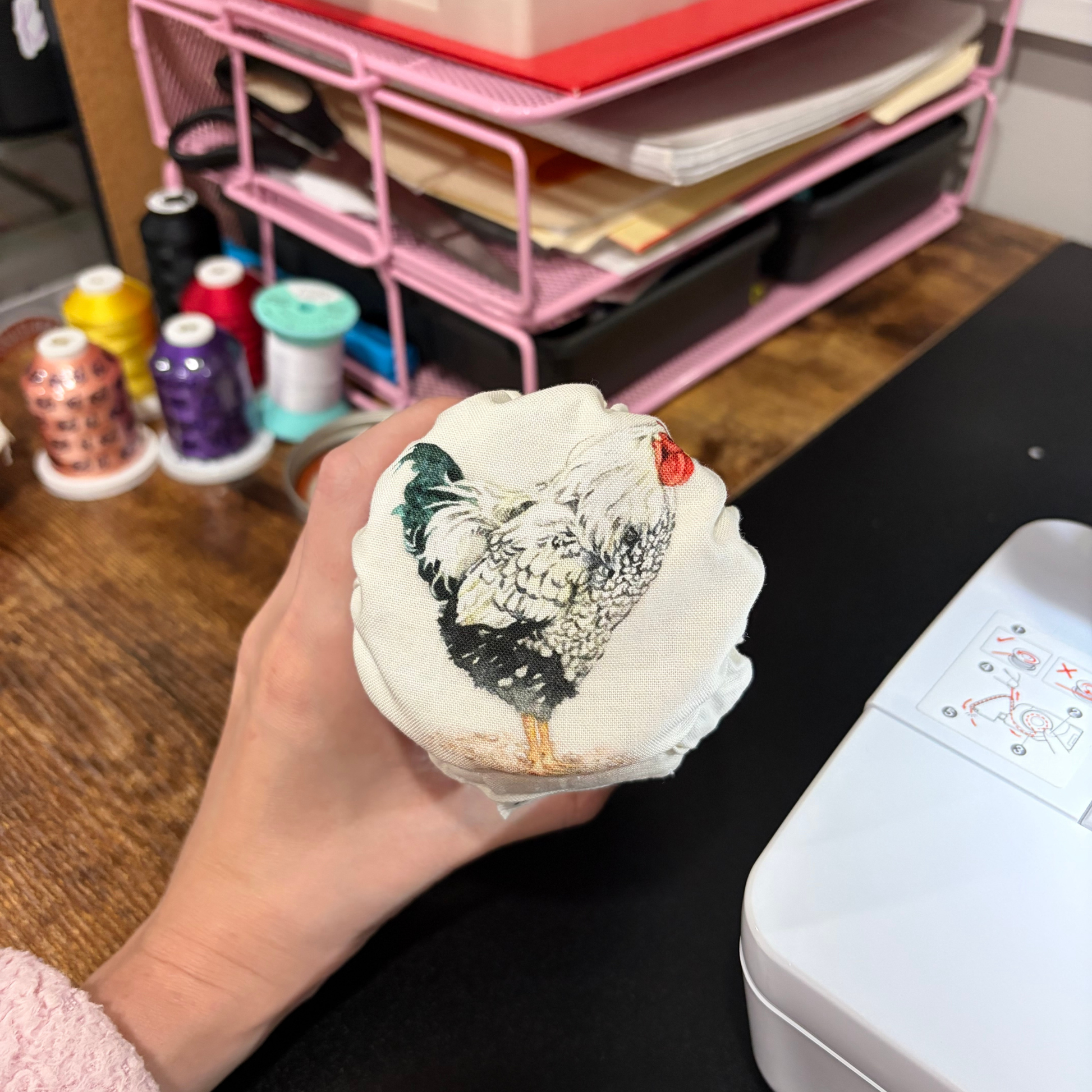 A hand holds a small jar with a cover featuring a printed rooster illustration, with a pink organizer, craft supplies, and a white box visible in the background.
