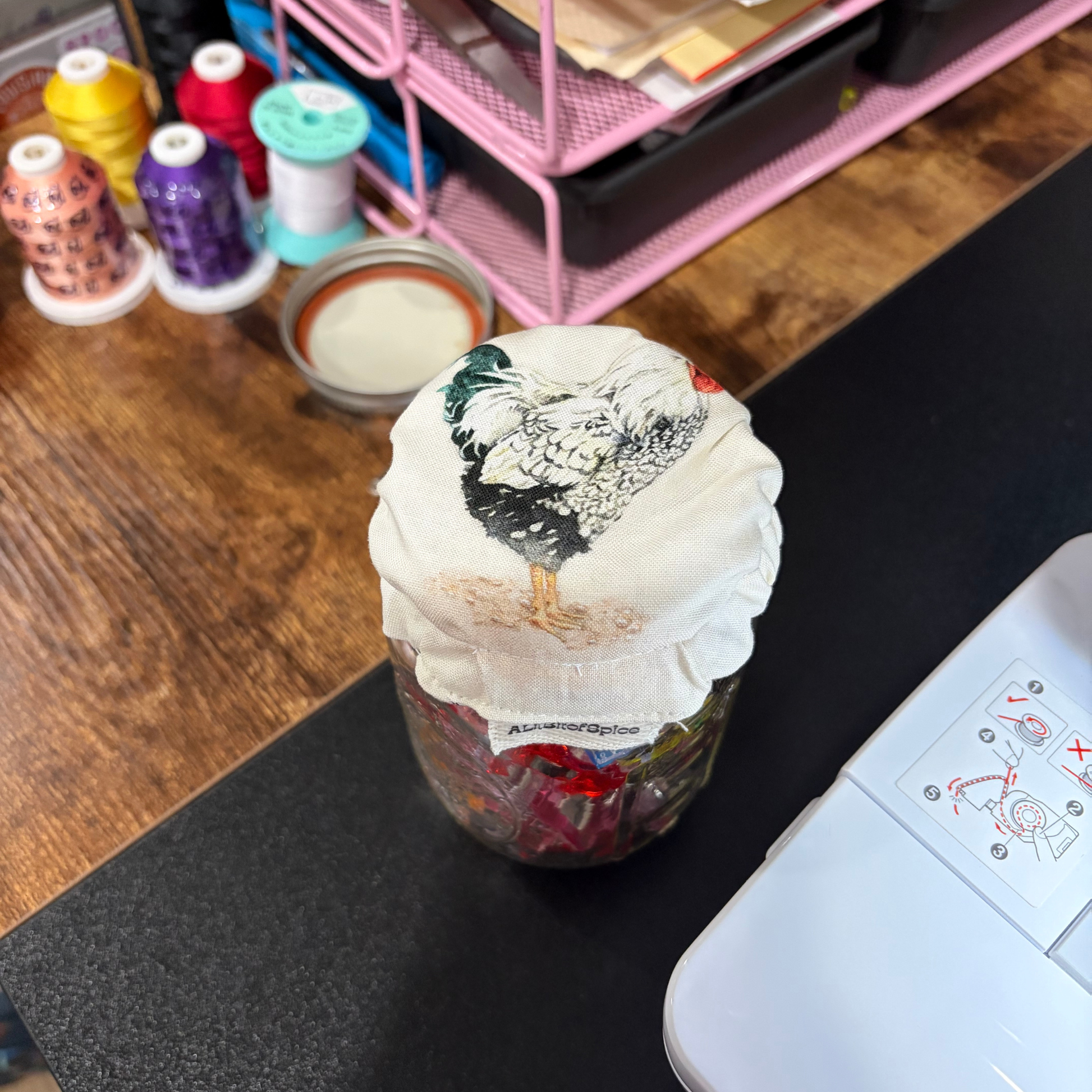 A clear glass jar filled with red and green items, covered with a white fabric lid printed with a black and white rooster, sitting on a black surface in a craft workspace with spools of thread and a pink organizer in the background.