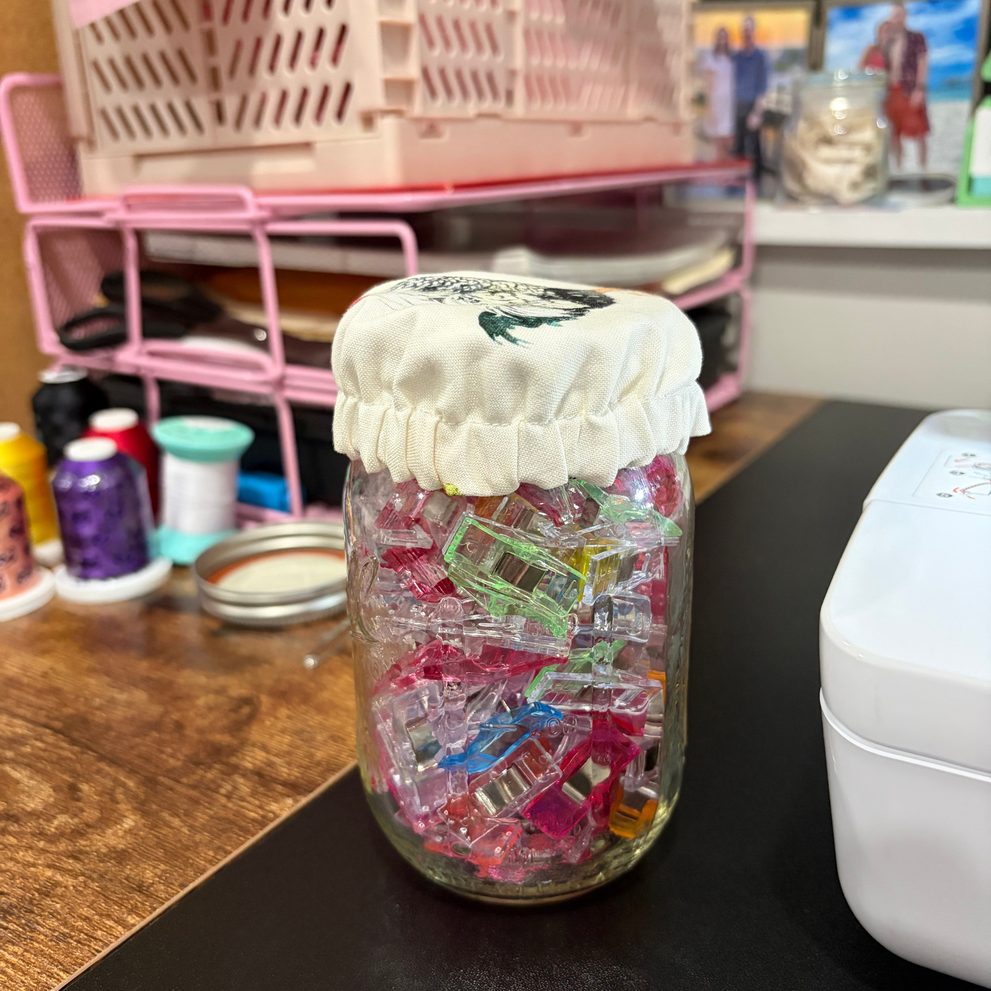 A clear glass jar filled with many colorful plastic clips or fasteners, covered with a white fabric lid, sitting on a dark tabletop in a craft or sewing workspace with spools of thread and pink storage shelves in the background.
