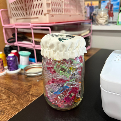 A clear glass jar filled with many colorful plastic clips or fasteners, covered with a white fabric lid, sitting on a dark tabletop in a craft or sewing workspace with spools of thread and pink storage shelves in the background.