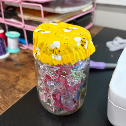 A clear glass jar filled with colorful clips, topped with a yellow fabric cover printed with white bees, set on a desk with craft supplies in the background.
