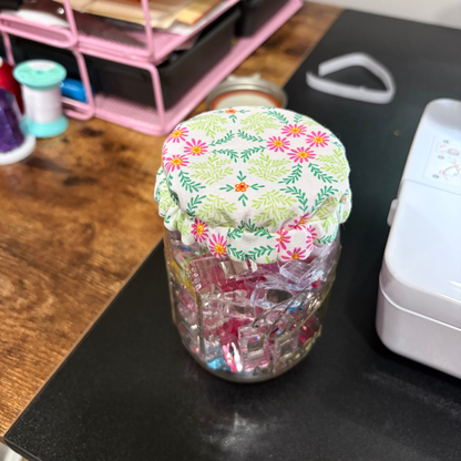 A clear glass jar filled with pink and blue sewing clips, topped with a fabric lid featuring a green and pink floral pattern, set on a black desk.