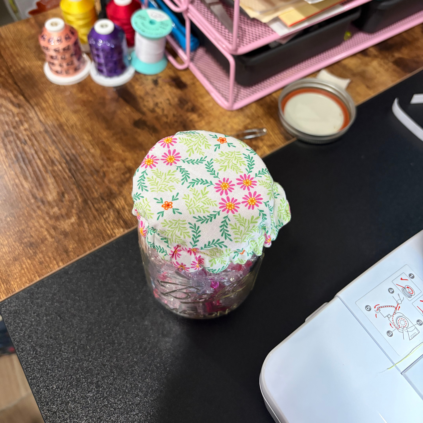 A clear glass jar covered with a light green fabric lid featuring a colorful floral and leaf print, placed on a black tabletop with sewing supplies in the background.