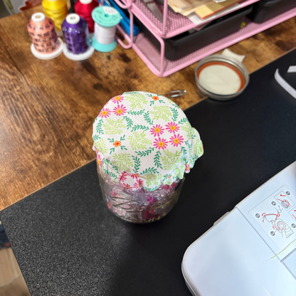 A clear glass jar covered with a light green fabric lid featuring a colorful floral and leaf print, placed on a black tabletop with sewing supplies in the background.
