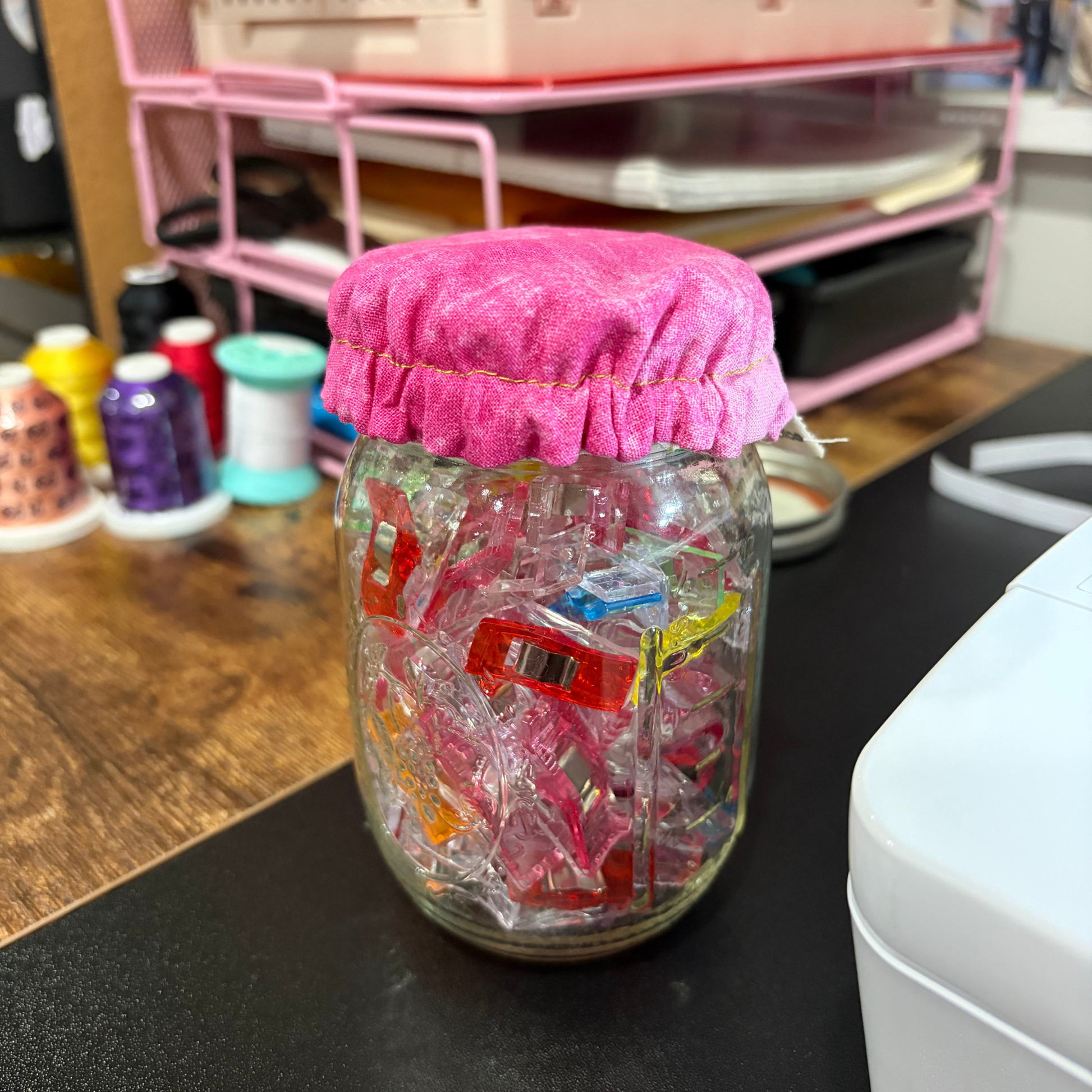 A clear glass mason jar filled with colorful plastic clips, sealed with a bright pink fabric cover, sitting on a dark tabletop in a craft room setting.