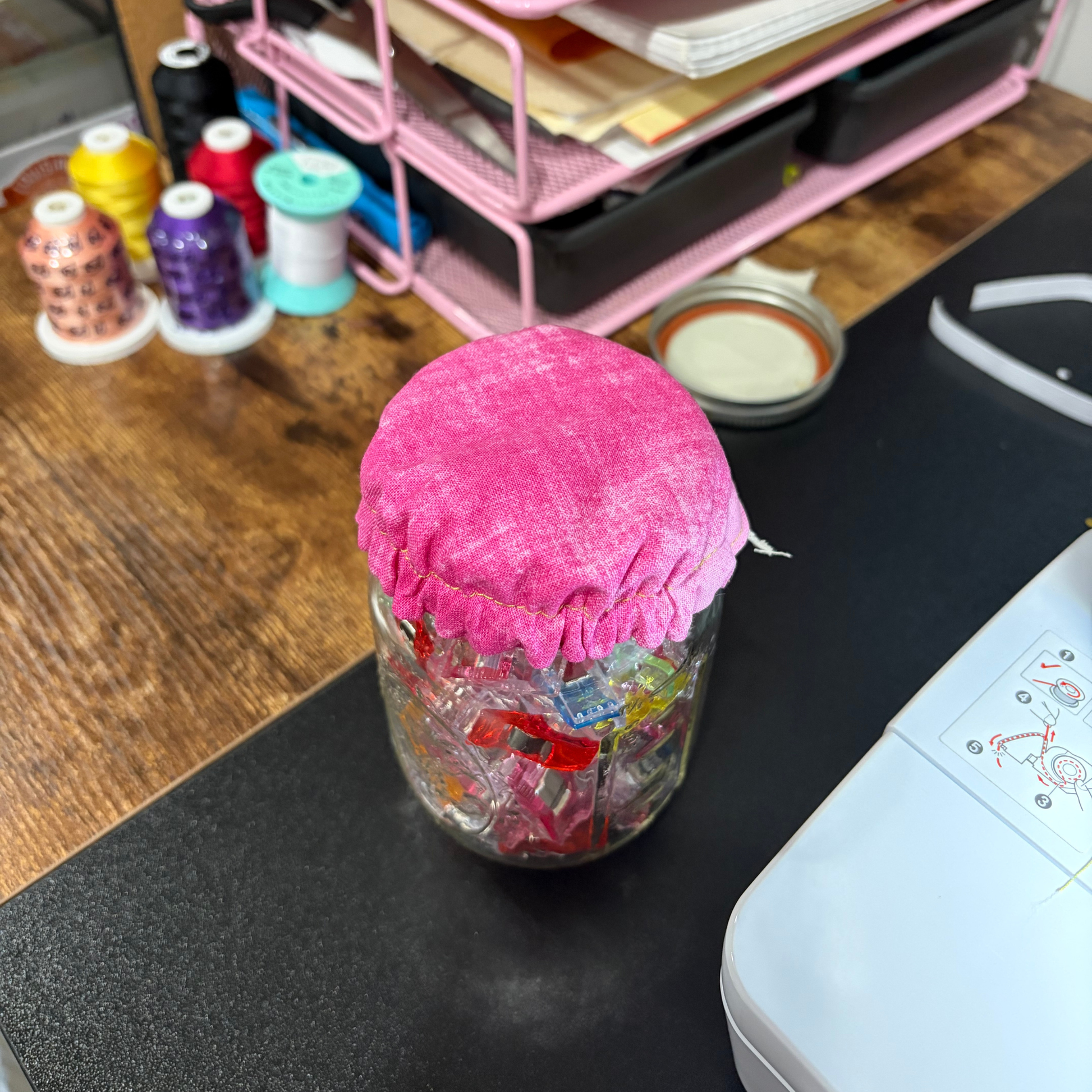 A clear glass jar filled with colorful craft ribbons or threads, covered with a bright pink fabric lid, sitting on a black desk mat in a sewing or crafting workspace.