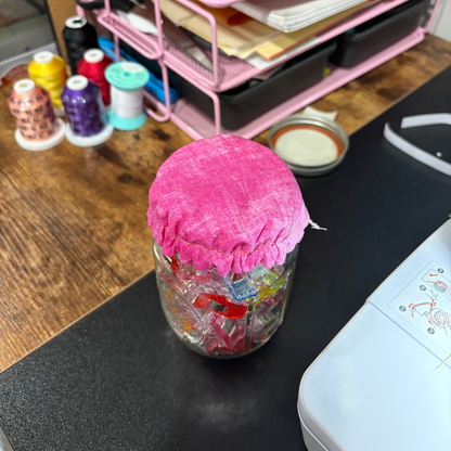 A clear glass jar filled with colorful craft ribbons or threads, covered with a bright pink fabric lid, sitting on a black desk mat in a sewing or crafting workspace.