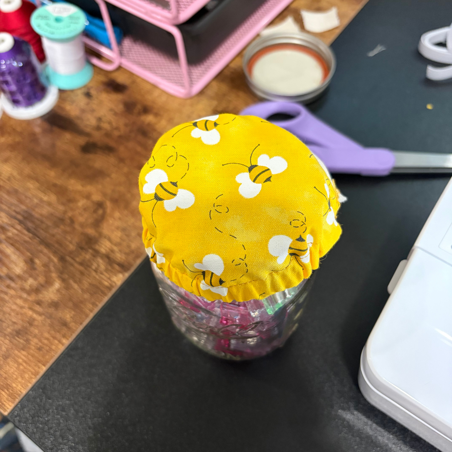 A clear glass jar filled with colorful items, covered at the top by a bright yellow fabric lid printed with white bees, set on a dark tabletop with craft supplies in the background.