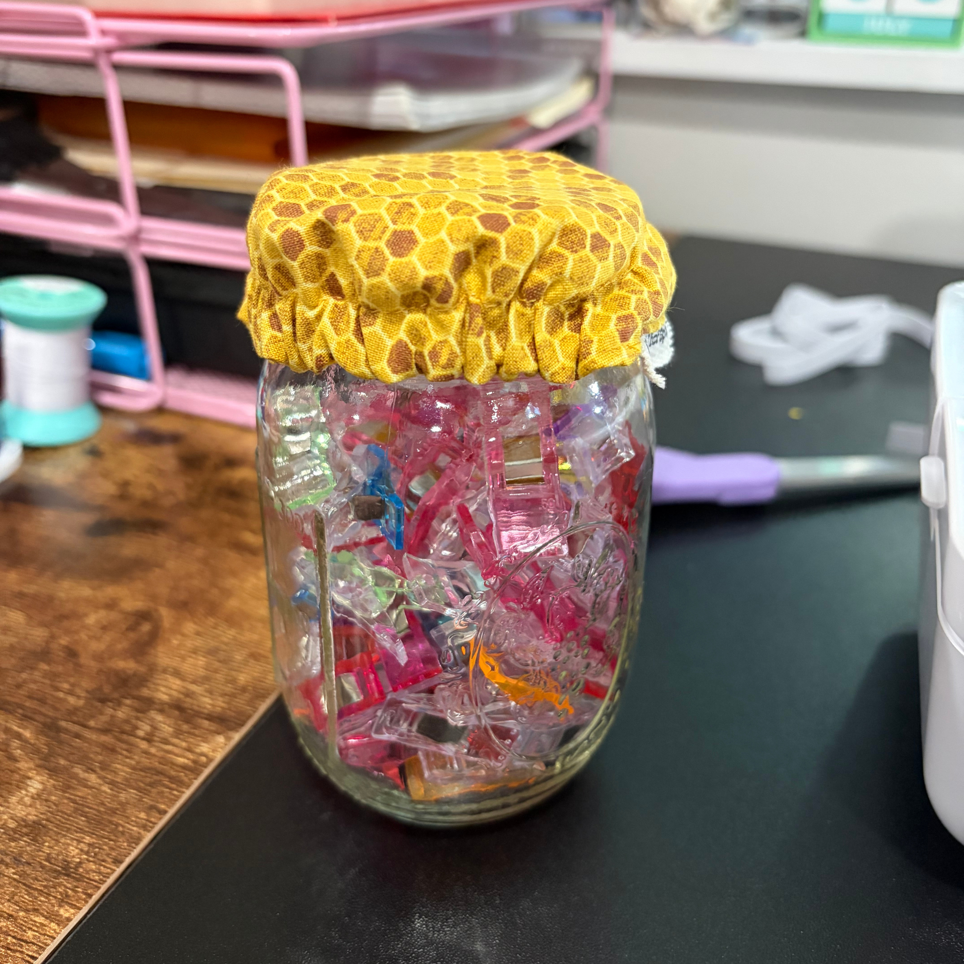 A clear glass jar filled with colorful plastic clips, topped with a yellow honeycomb-patterned fabric cover, sitting on a dark tabletop in a craft room setting.