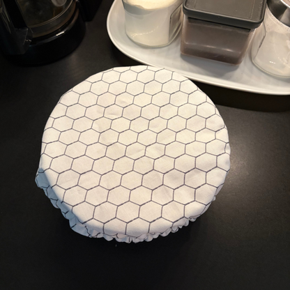 A round white fabric cover with a black hexagonal chicken wire pattern and a ruffled edge, shown on a dark countertop with kitchen items in the background.