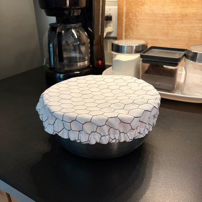 A round bowl covered with a white fabric lid featuring a black chicken wire pattern and ruffled edges, sitting on a dark kitchen countertop with a coffee maker and storage containers in the background.
