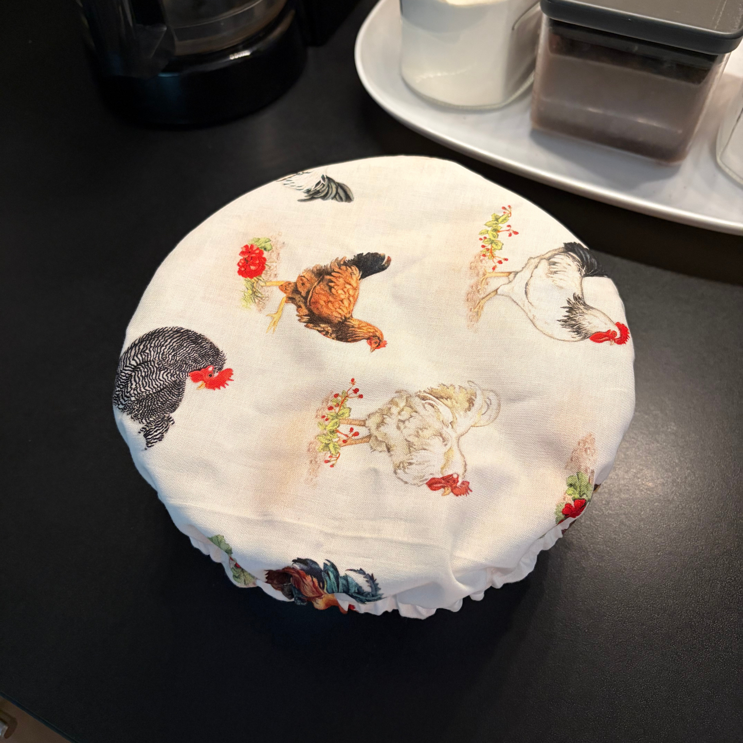 A round white fabric cover with a printed chicken pattern, draped over a round container on a dark tabletop.