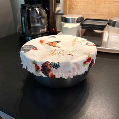 A round bowl covered with a white fabric lid printed with colorful rooster and chicken illustrations, sitting on a dark kitchen countertop.