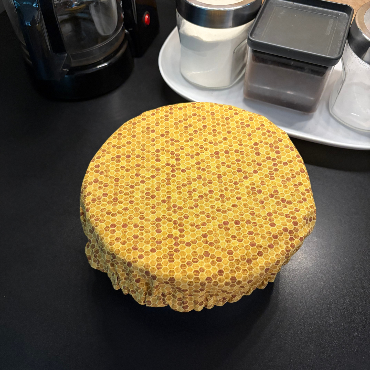 A round fabric cover with a yellow and brown honeycomb pattern, draped over a round container on a dark countertop with kitchen items in the background.