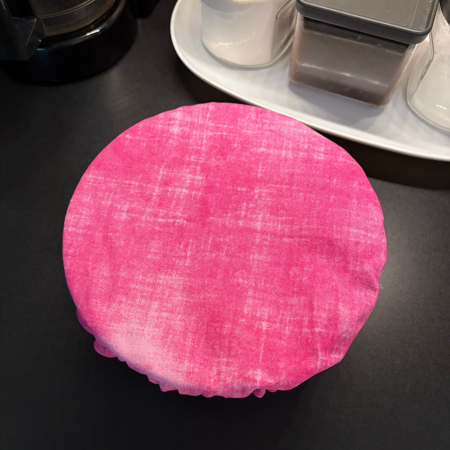A round pink fabric cover with a textured linen-like weave, shown draped over a round container on a dark countertop with kitchen items in the background.