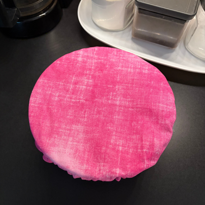 A round pink fabric cover with a textured linen-like weave, shown draped over a round container on a dark countertop with kitchen items in the background.