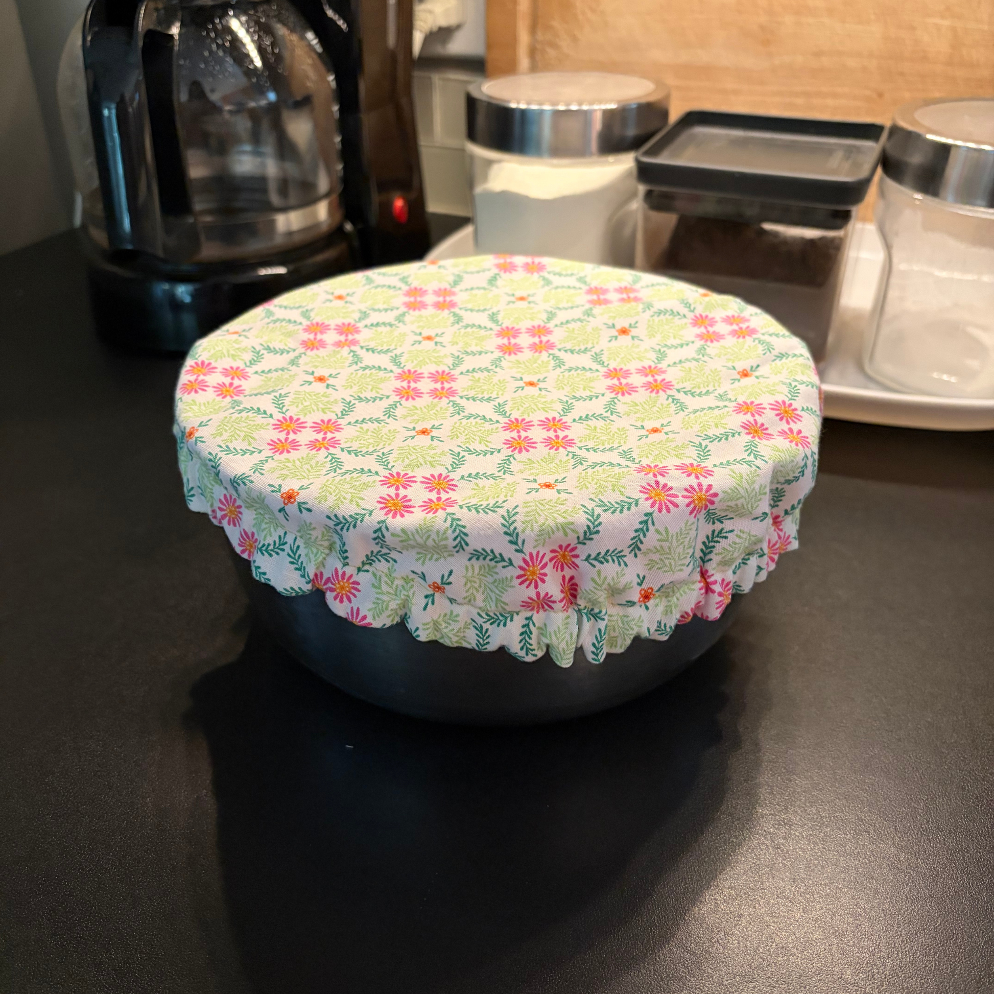 A round kitchen container covered with a light green fabric lid featuring a pink and green floral pattern and a ruffled edge, sitting on a dark countertop in a kitchen setting.