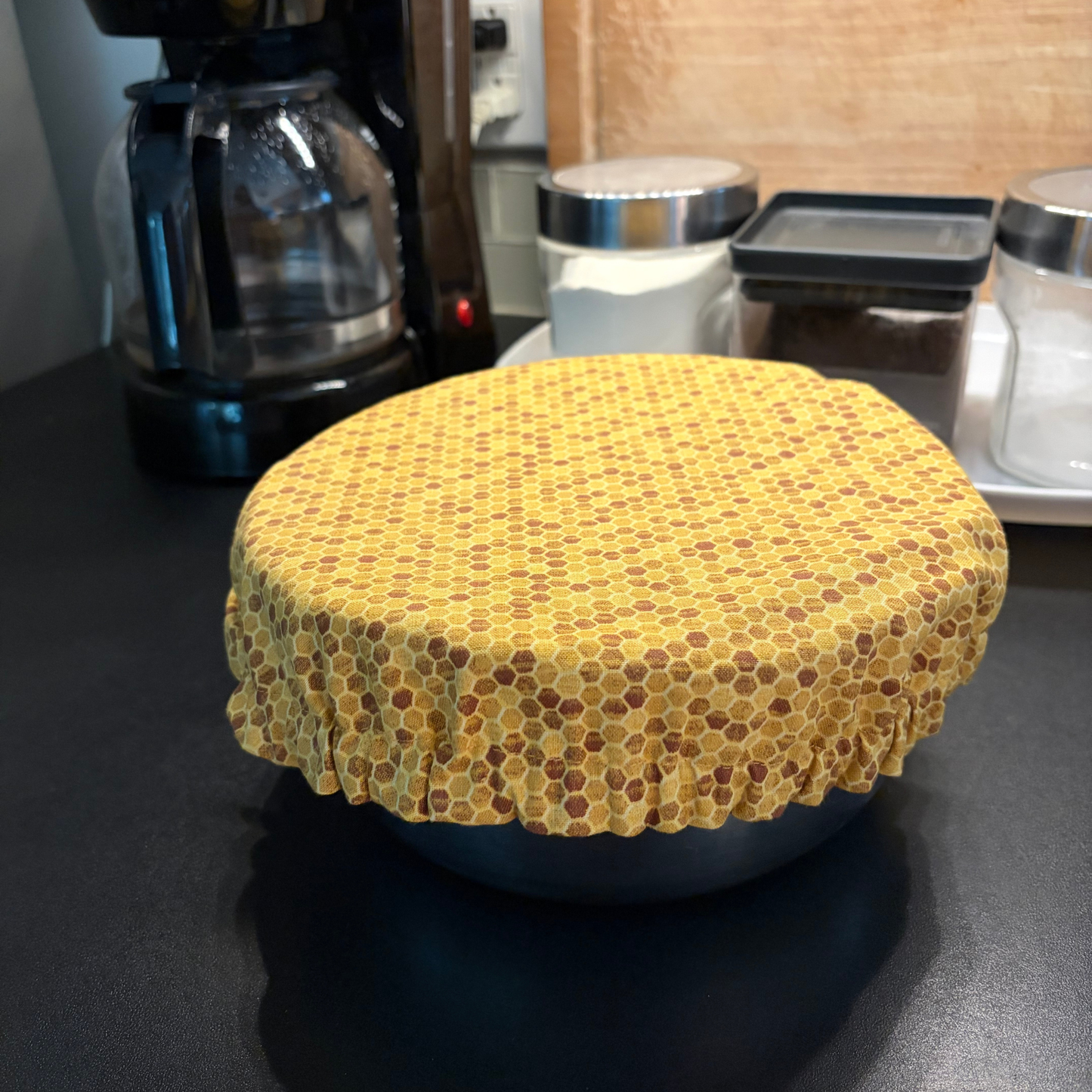 A round bowl covered with a yellow honeycomb-patterned fabric, placed on a dark kitchen counter with a coffee maker and storage containers in the background.
