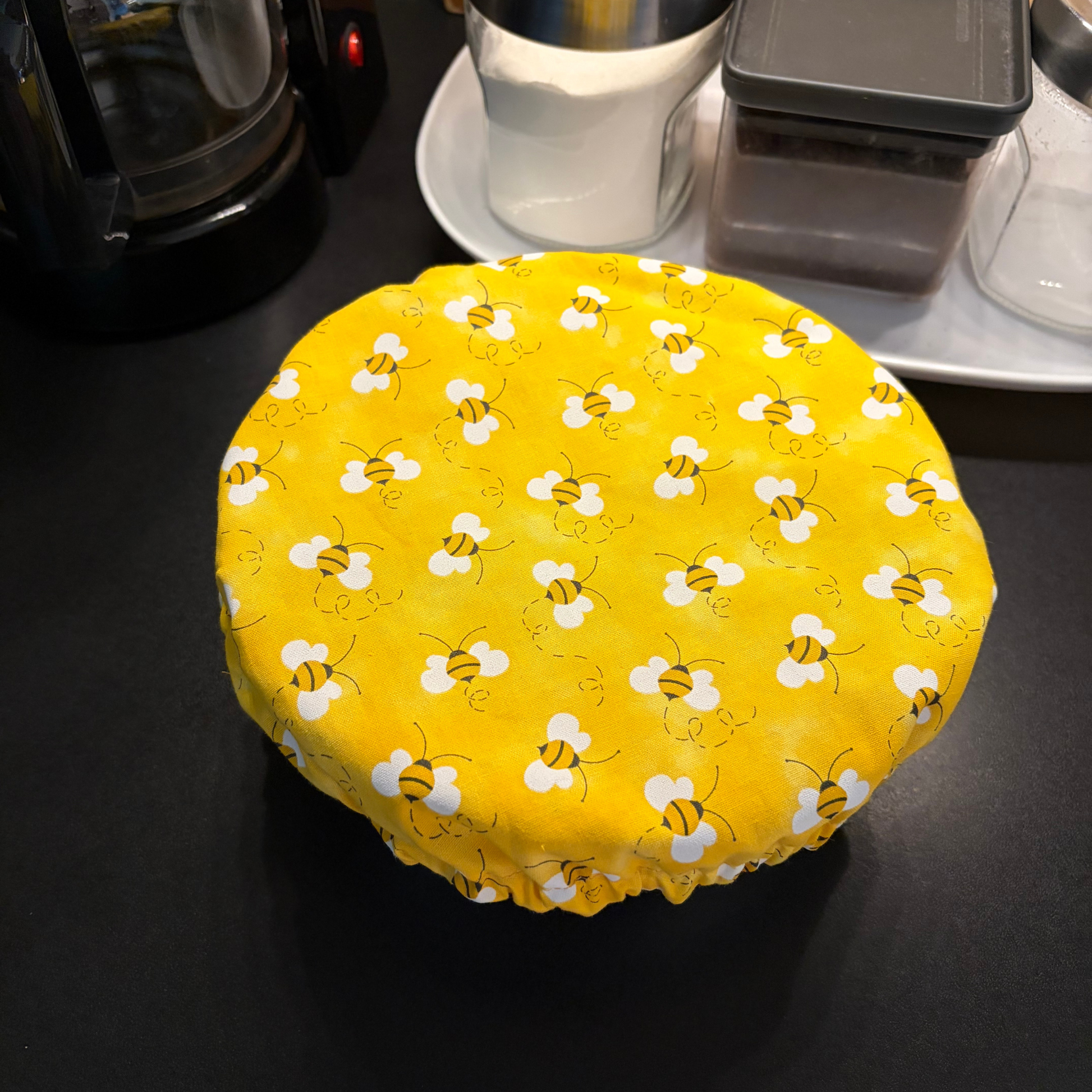 A round fabric bowl cover with a yellow background and a repeating white bee pattern, shown wrapped around a round container on a dark countertop.