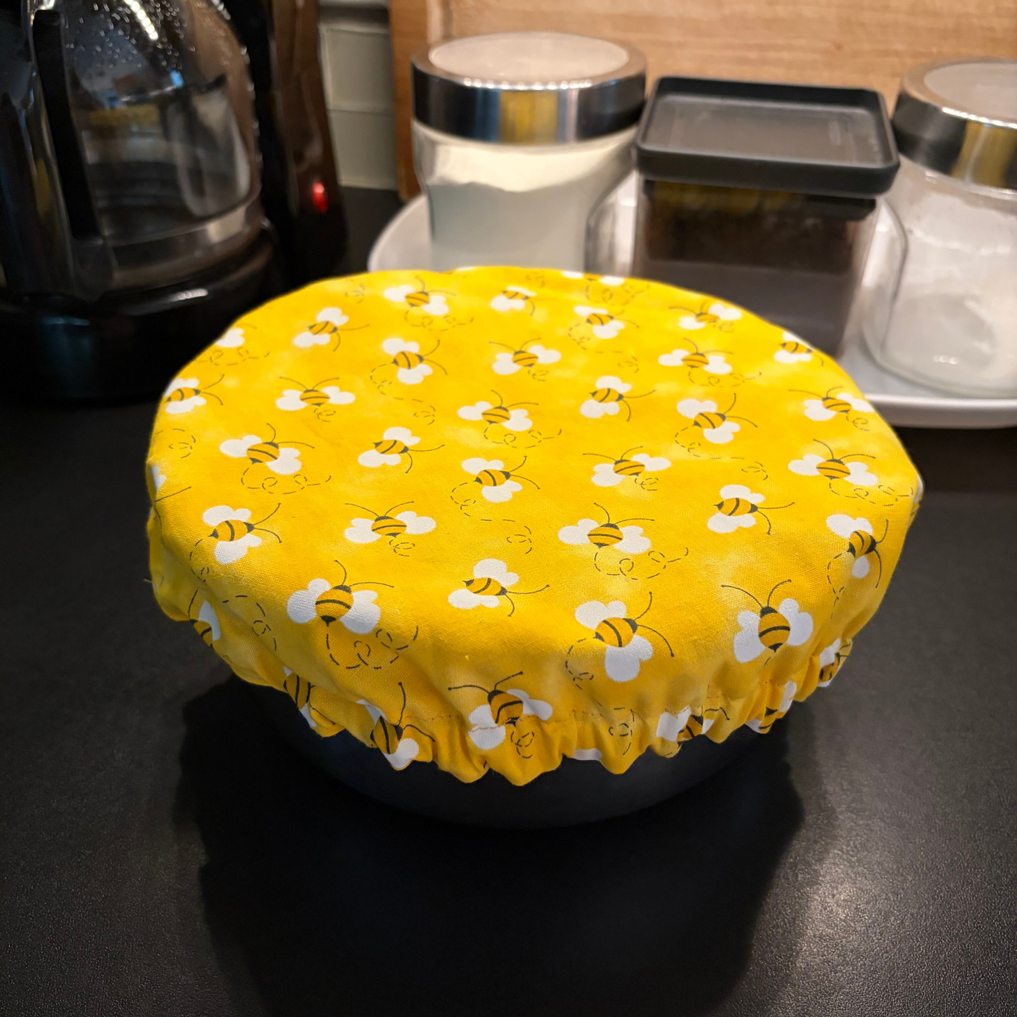 A round yellow fabric cover with a repeating white bee pattern, stretched over a dark bowl on a dark countertop in a kitchen setting.