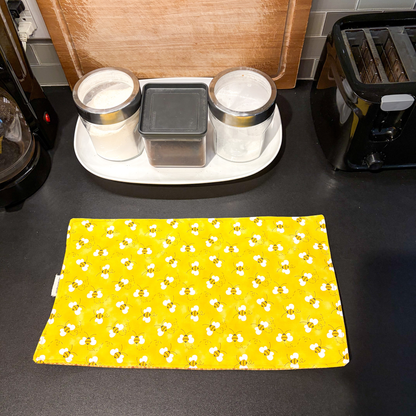 A yellow fabric placemat with a bumblebee pattern is laid out on a dark kitchen countertop, with a white tray of containers, a wooden cutting board, and a black toaster in the background.