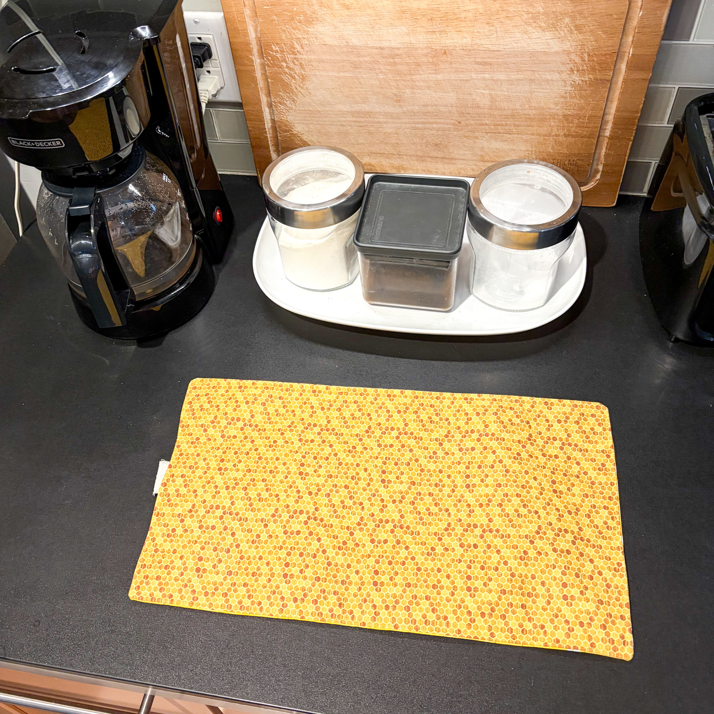 A rectangular yellow kitchen mat with a small honeycomb pattern lies on a dark countertop in a kitchen setting, with a coffee maker, canisters, and a wooden cutting board in the background.