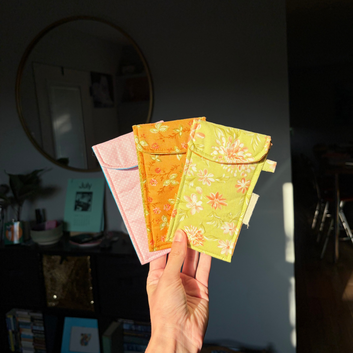 A hand holds three small pouches in pink, orange, and yellow-green floral prints, photographed in a real indoor room with sunlight and a mirror in the background.