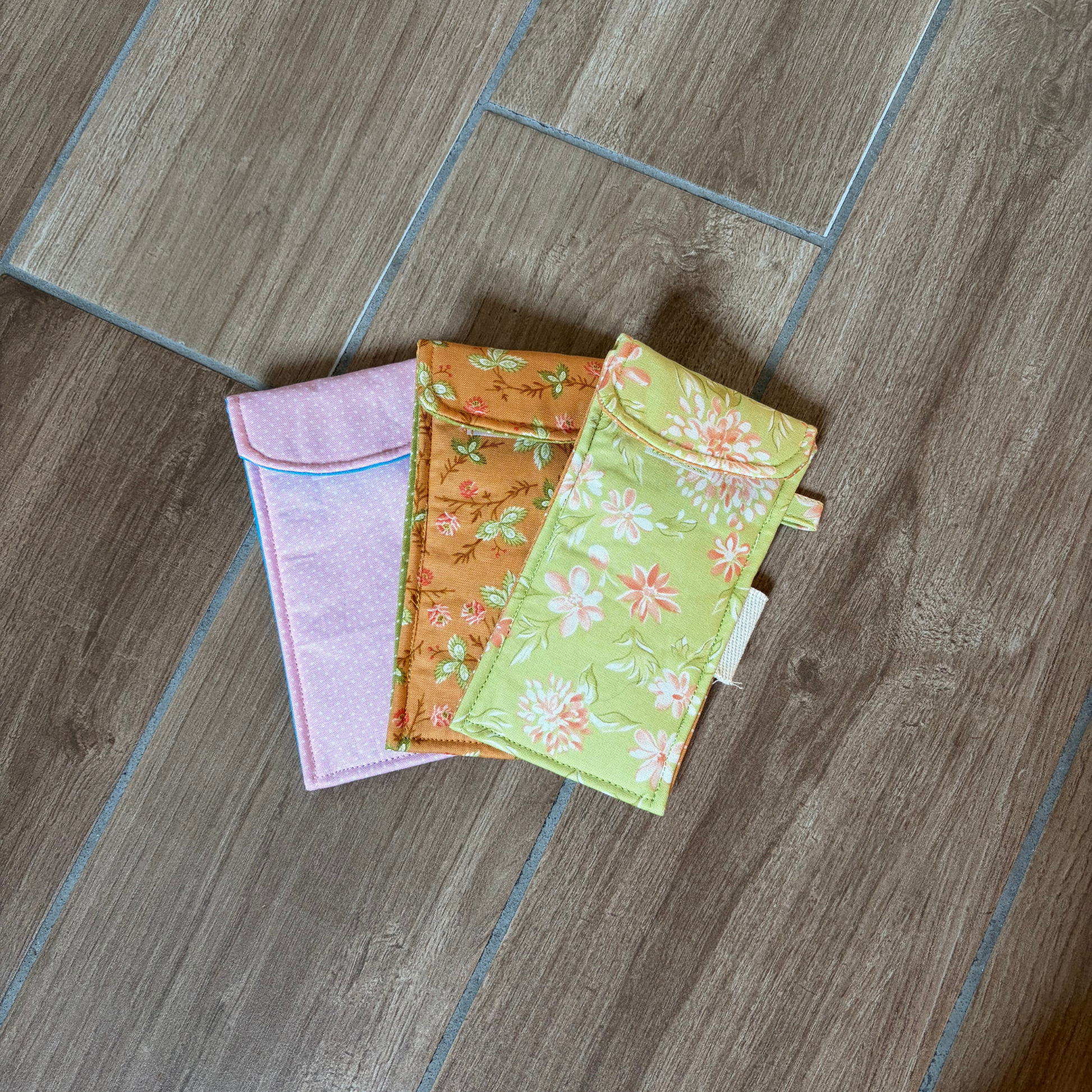 Three small fabric pouches with floral prints are arranged on a wood floor. One is light pink with a blue border, one is orange with a floral pattern, and one is light green with a white floral print.